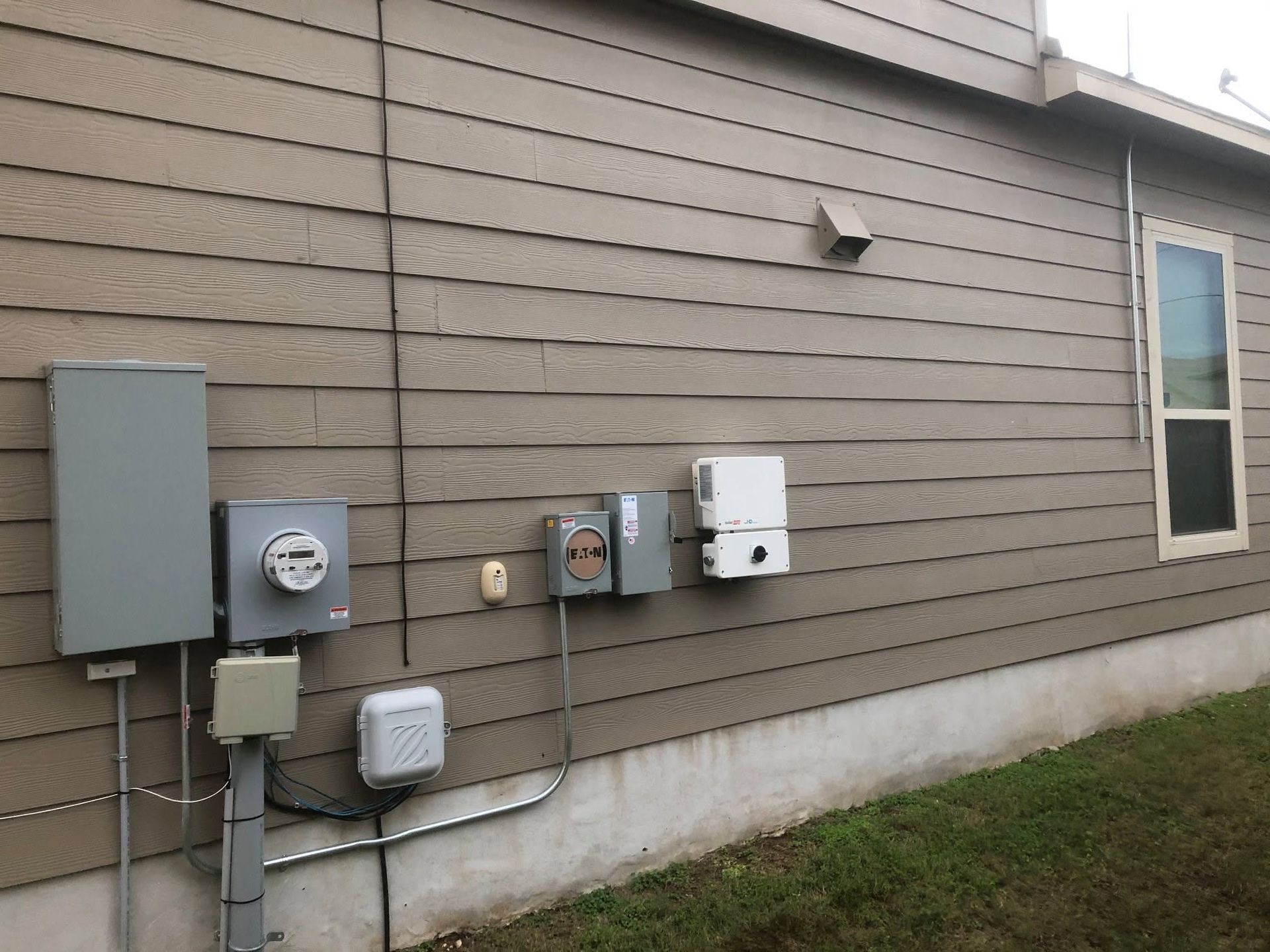 Exterior house wall with several utility meters and electrical boxes mounted on beige siding.