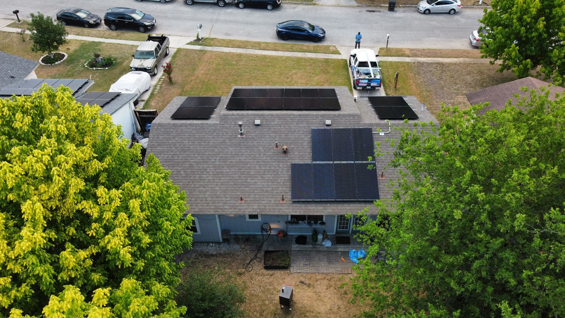 Aerial view of a house roof with solar panels, a large tree, and parked cars along the street.