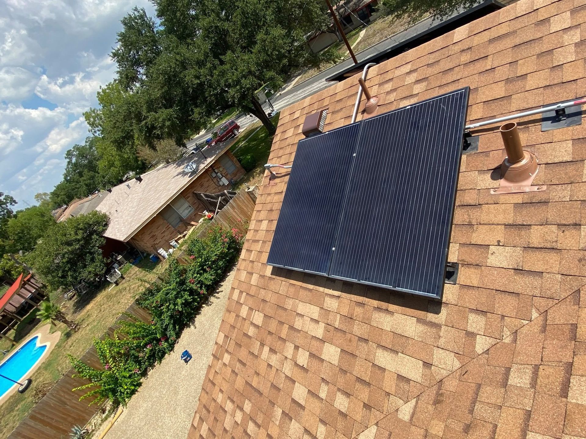 Tilted rooftop with a dark solar panel mounted on tan shingles under a partly cloudy sky