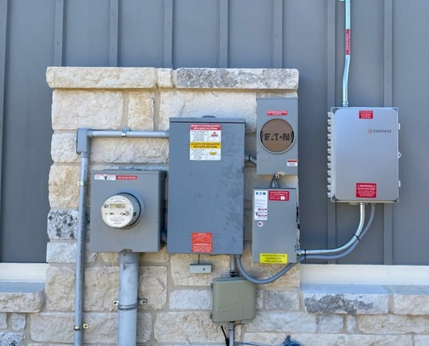 Electrical meters and utility boxes mounted on a stone wall beside a gray building.