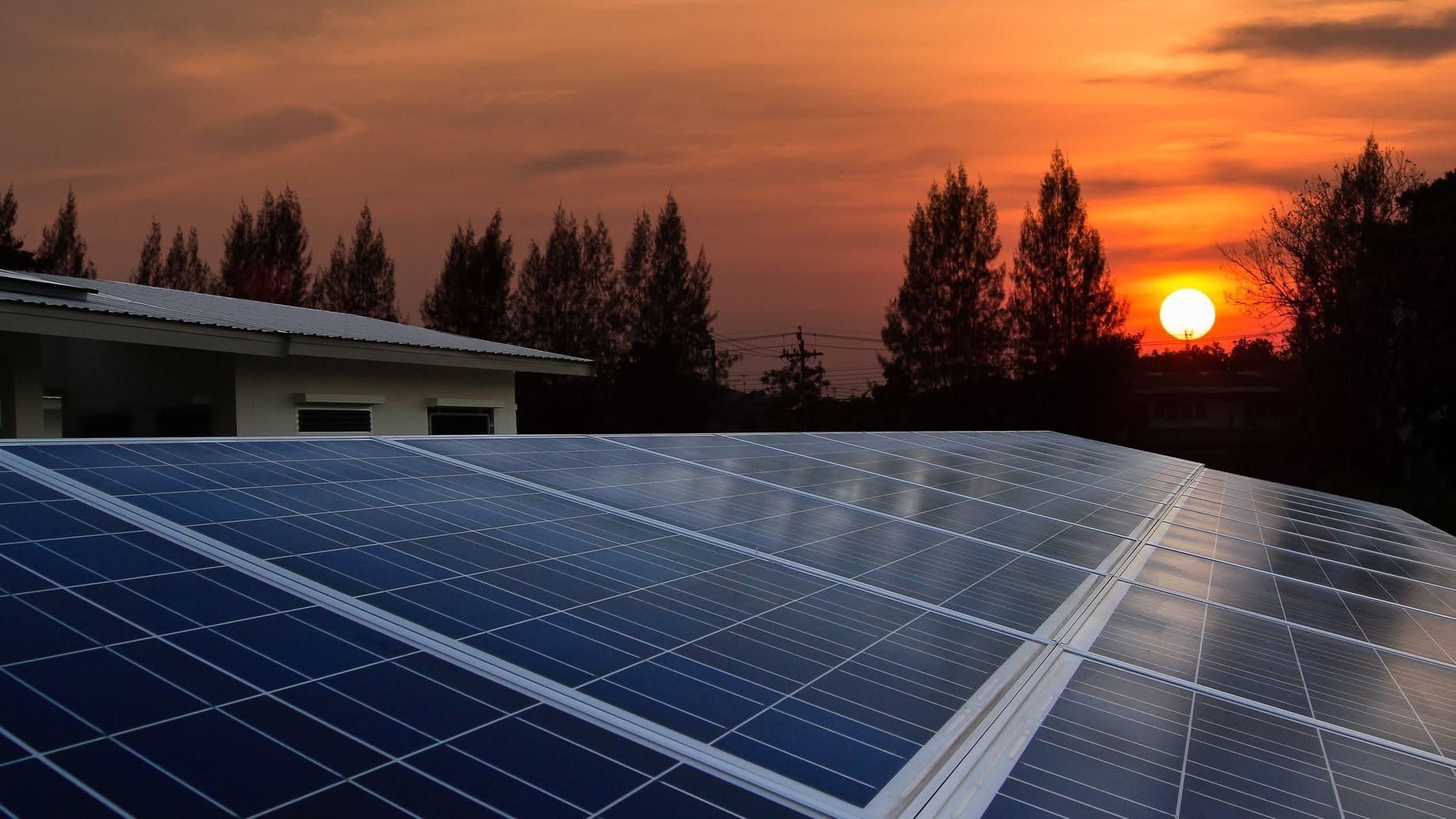 Solar panels on a rooftop at sunset with an orange sky and silhouetted trees in the background