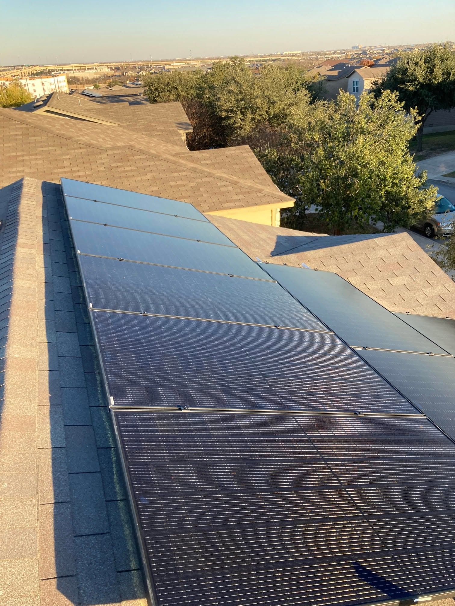 Solar panels on a rooftop beside tan shingles, with trees and a sunny suburban view in the background.