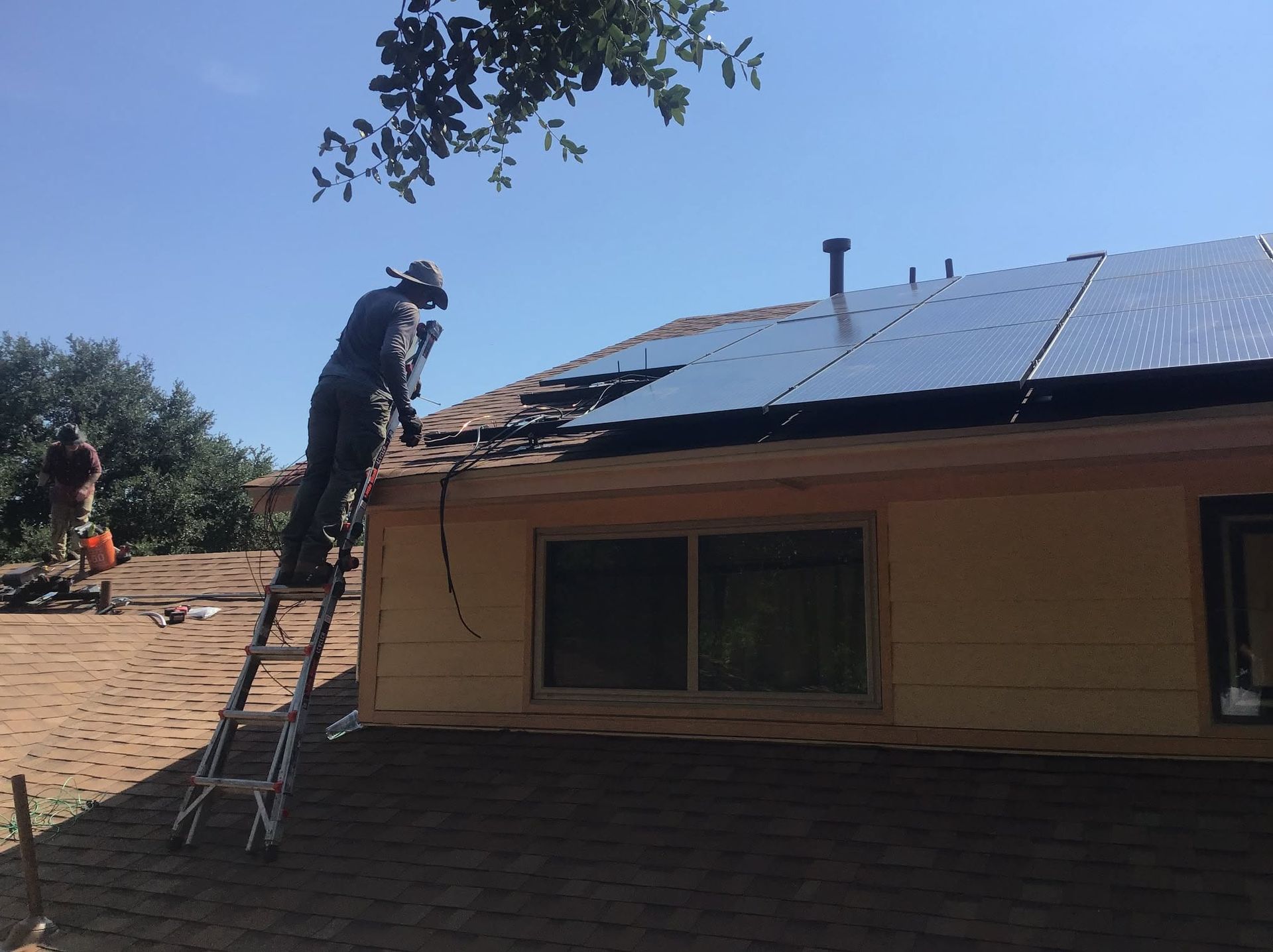 Worker on a ladder installing solar panels on a house roof under a clear blue sky.