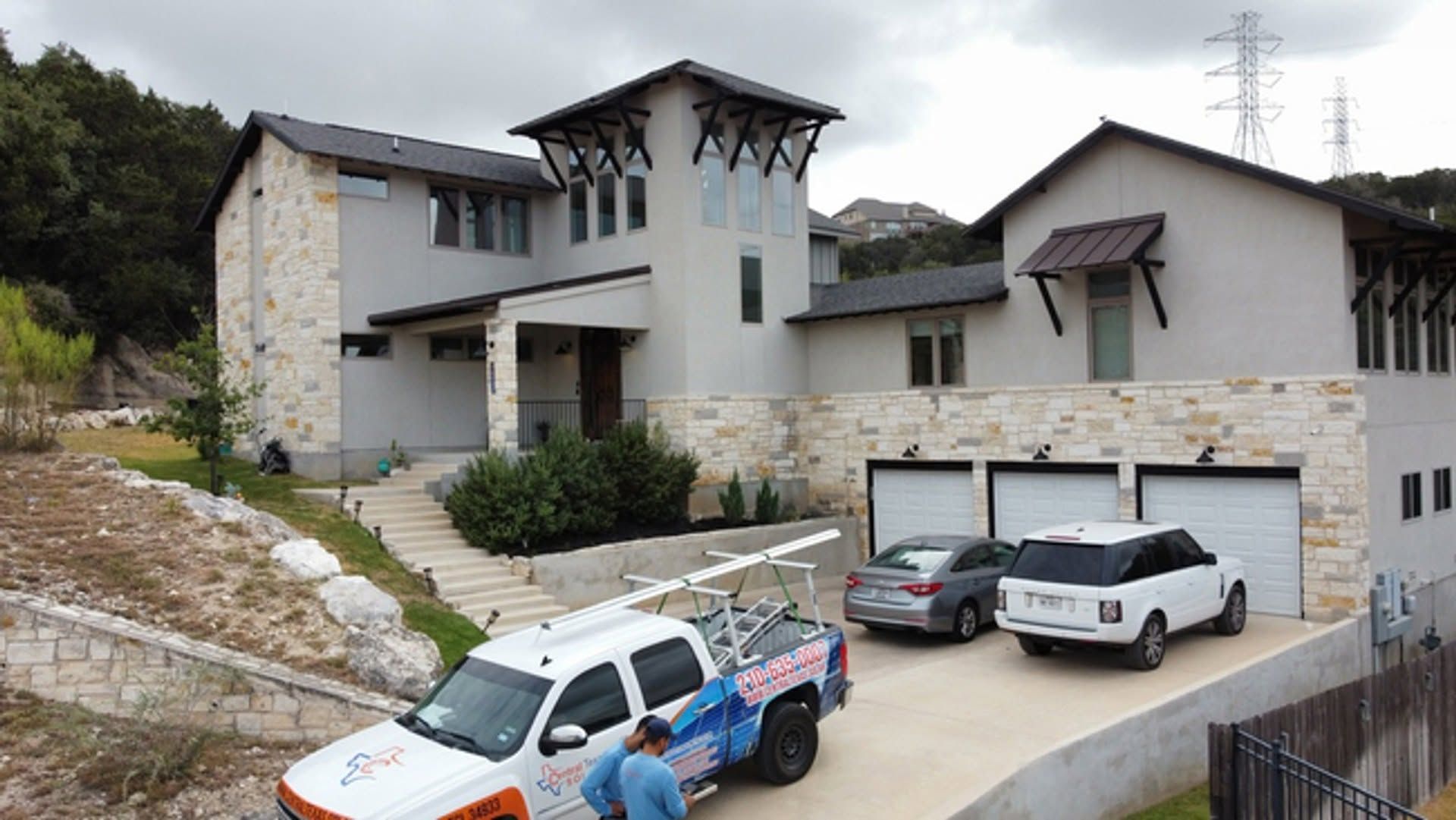 Modern stone house with three cars in a sloped driveway and a truck parked at the curb.