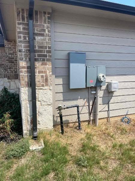 Utility meters and electrical boxes mounted on the side of a brick house above dry grass.
