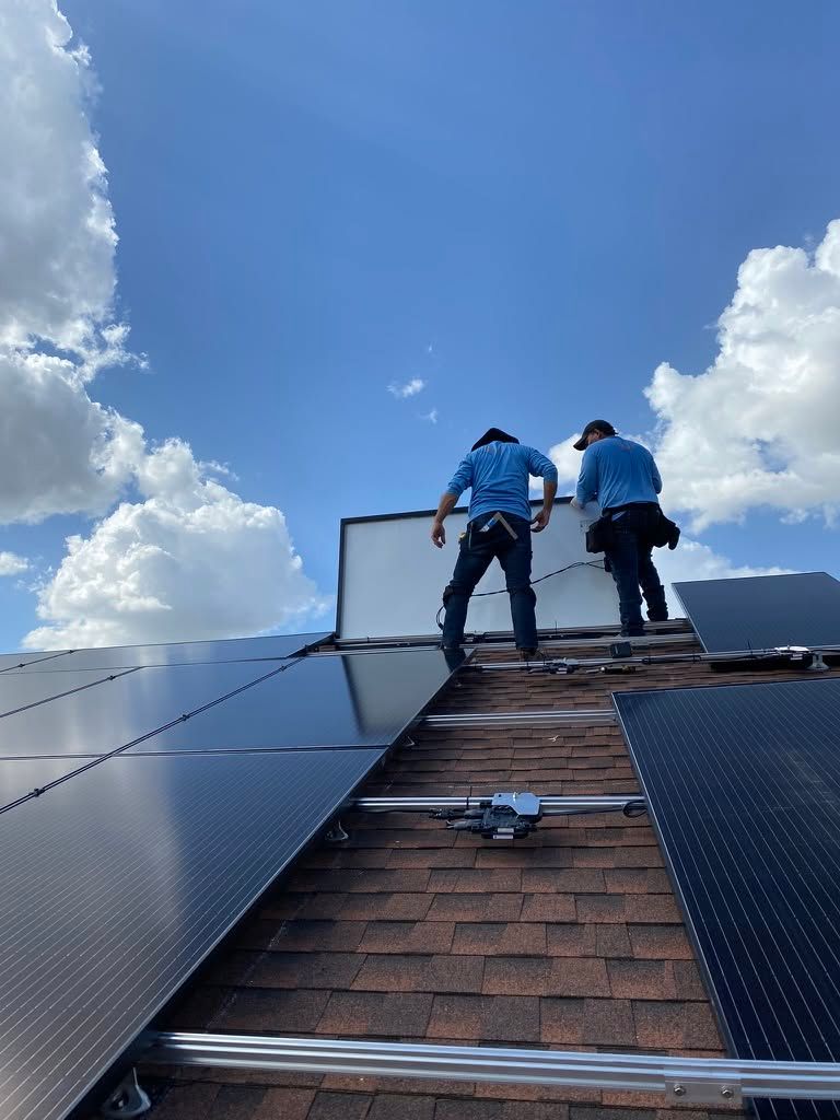 Two workers on a steep roof installing solar panels under a bright blue sky with clouds