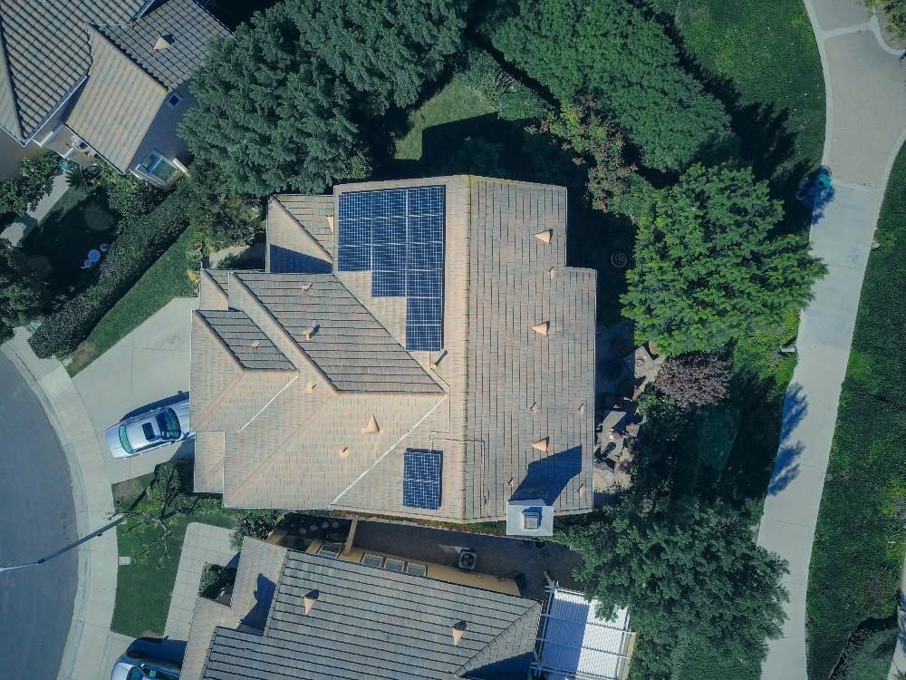 Aerial view of two suburban houses with tan roofs and solar panels, surrounded by trees.