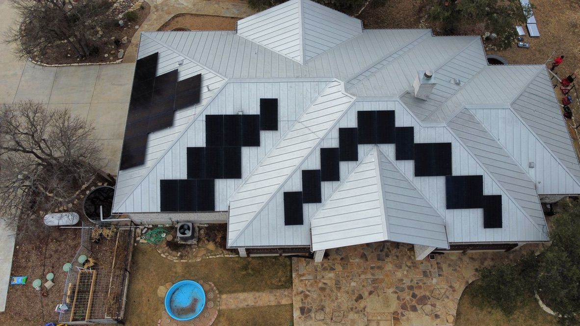 Aerial view of a house with a white roof and black geometric panels, surrounded by a yard and trees