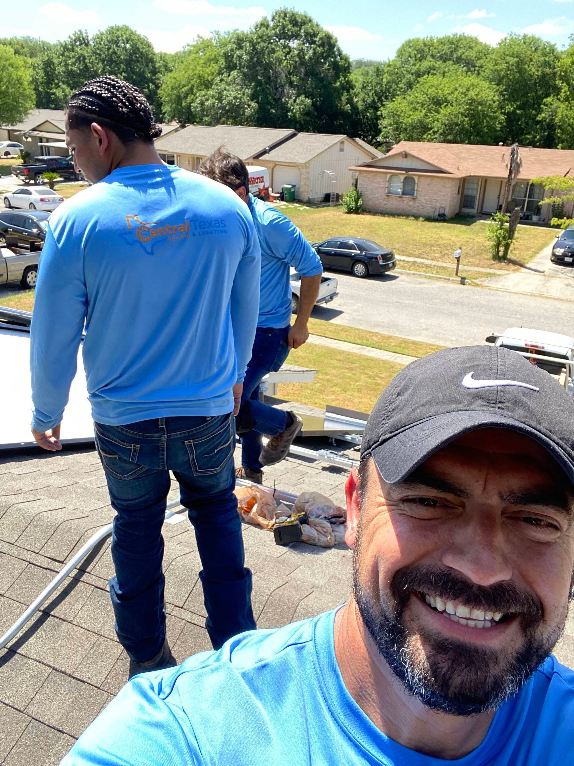 Two men on a rooftop in blue shirts, one smiling at the camera, with a suburban street below.