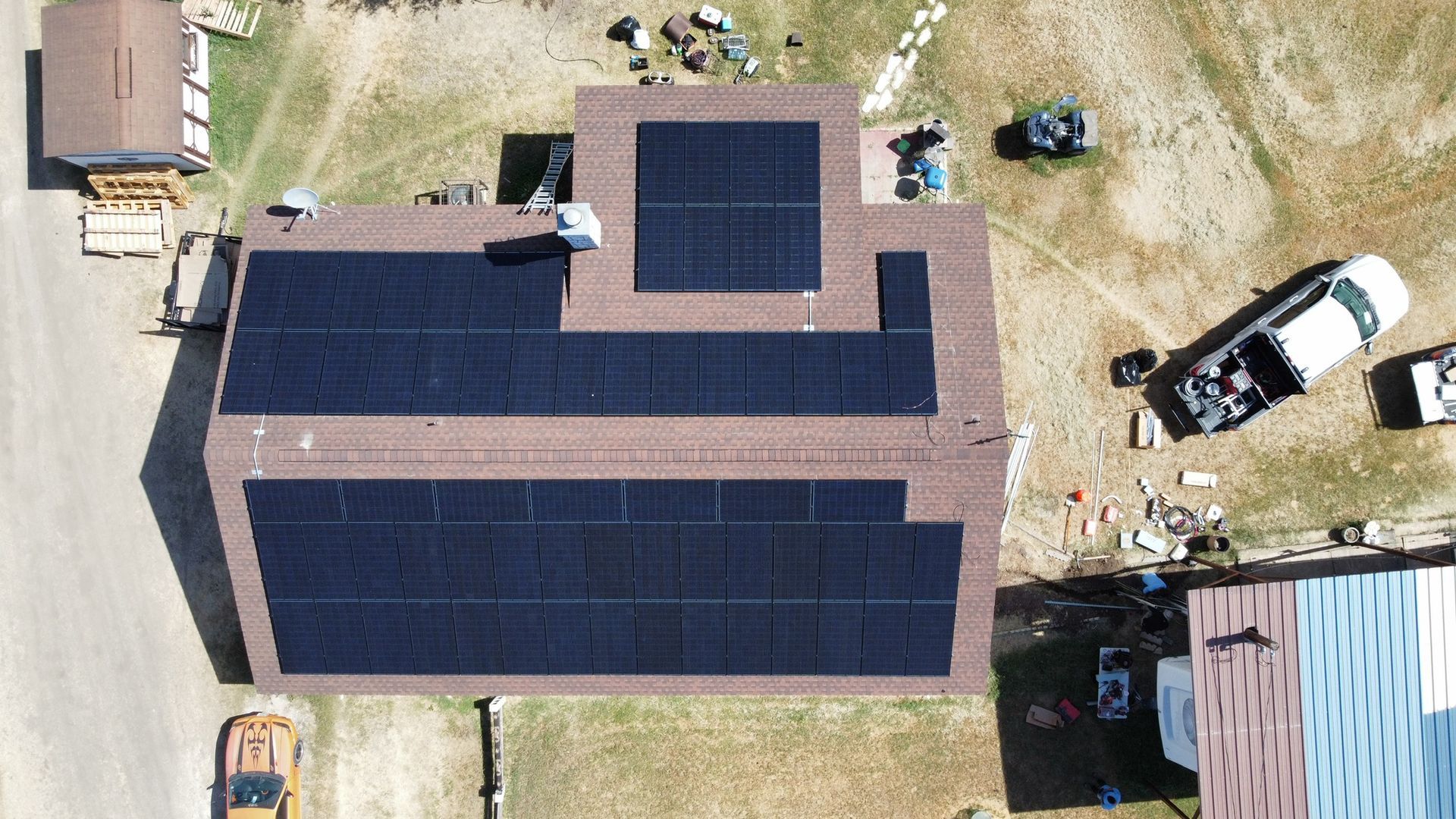 Aerial view of a house with large solar panel arrays on the roof and surrounding yard equipment.