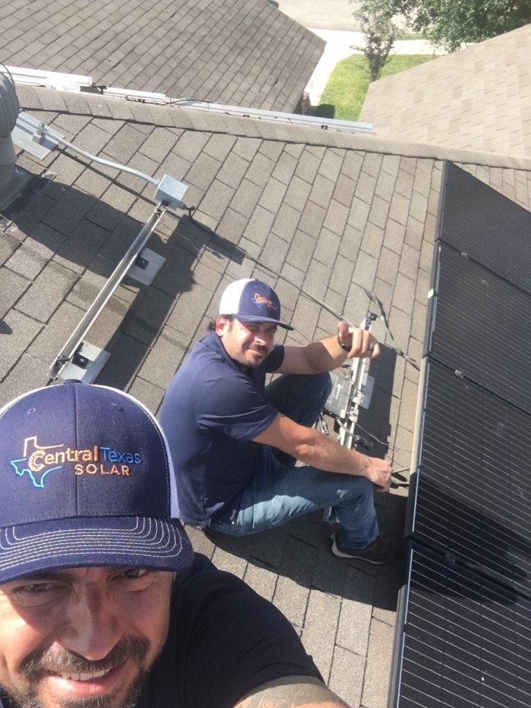 Two rooftop solar workers installing panels on a shingled roof, one smiling at the camera.