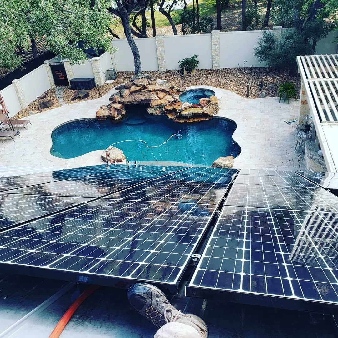 Backyard pool with solar panels in the foreground and rocks, trees, and a white fence behind
