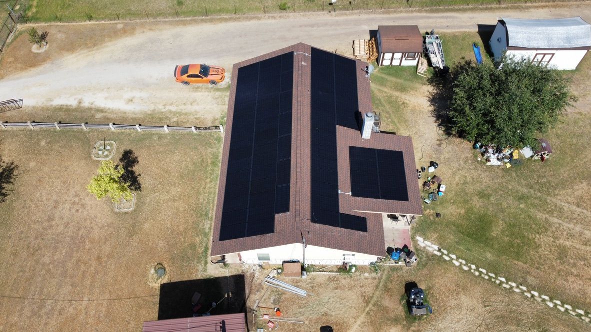 Aerial view of a house with black solar panels on the roof, a driveway, and a parked orange car.