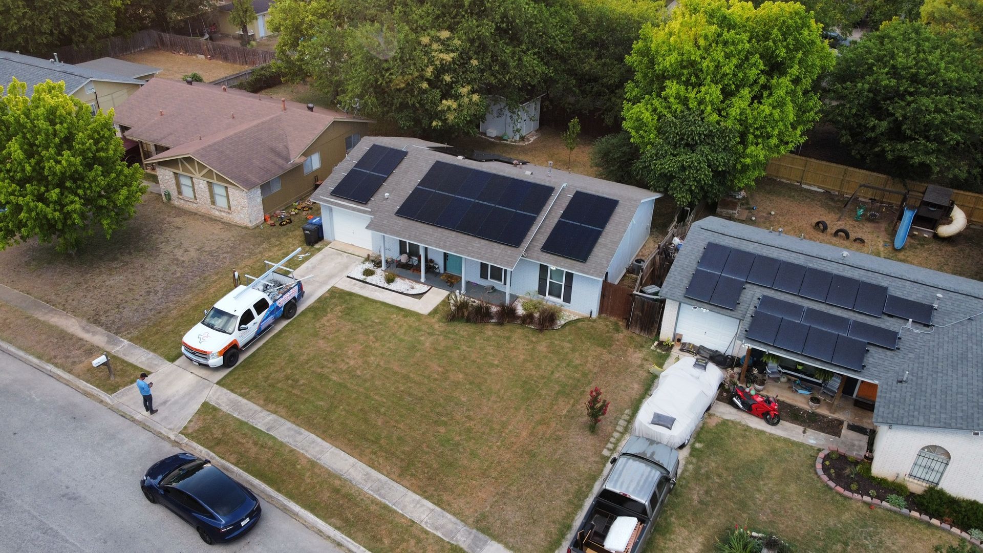 Aerial view of two suburban houses with solar panels, parked vehicles, and a grassy front yard.