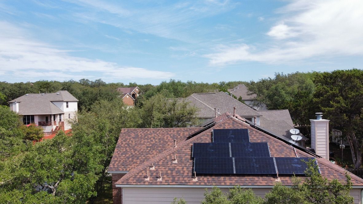 Suburban rooftops with solar panels under a blue sky, surrounded by green trees