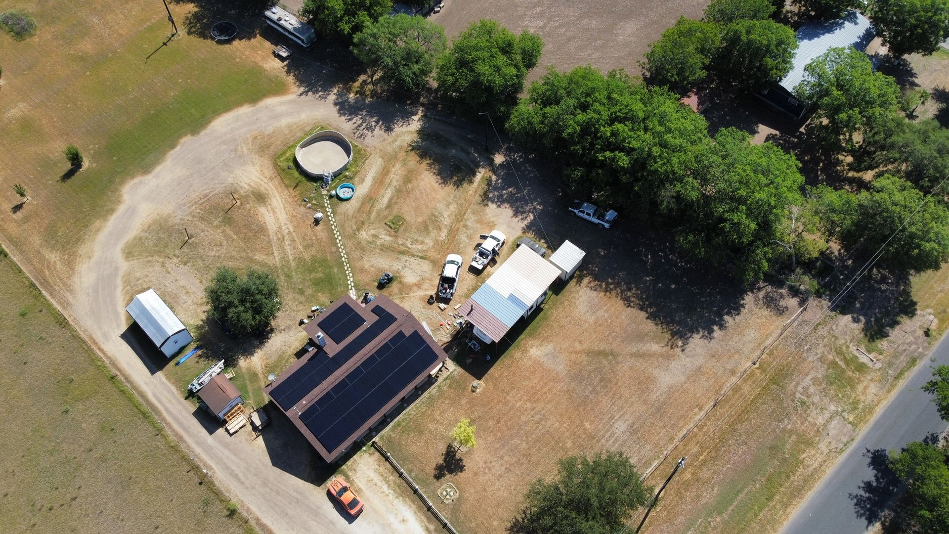 Aerial view of a rural property with a house, sheds, parked vehicles, and surrounding trees and open land