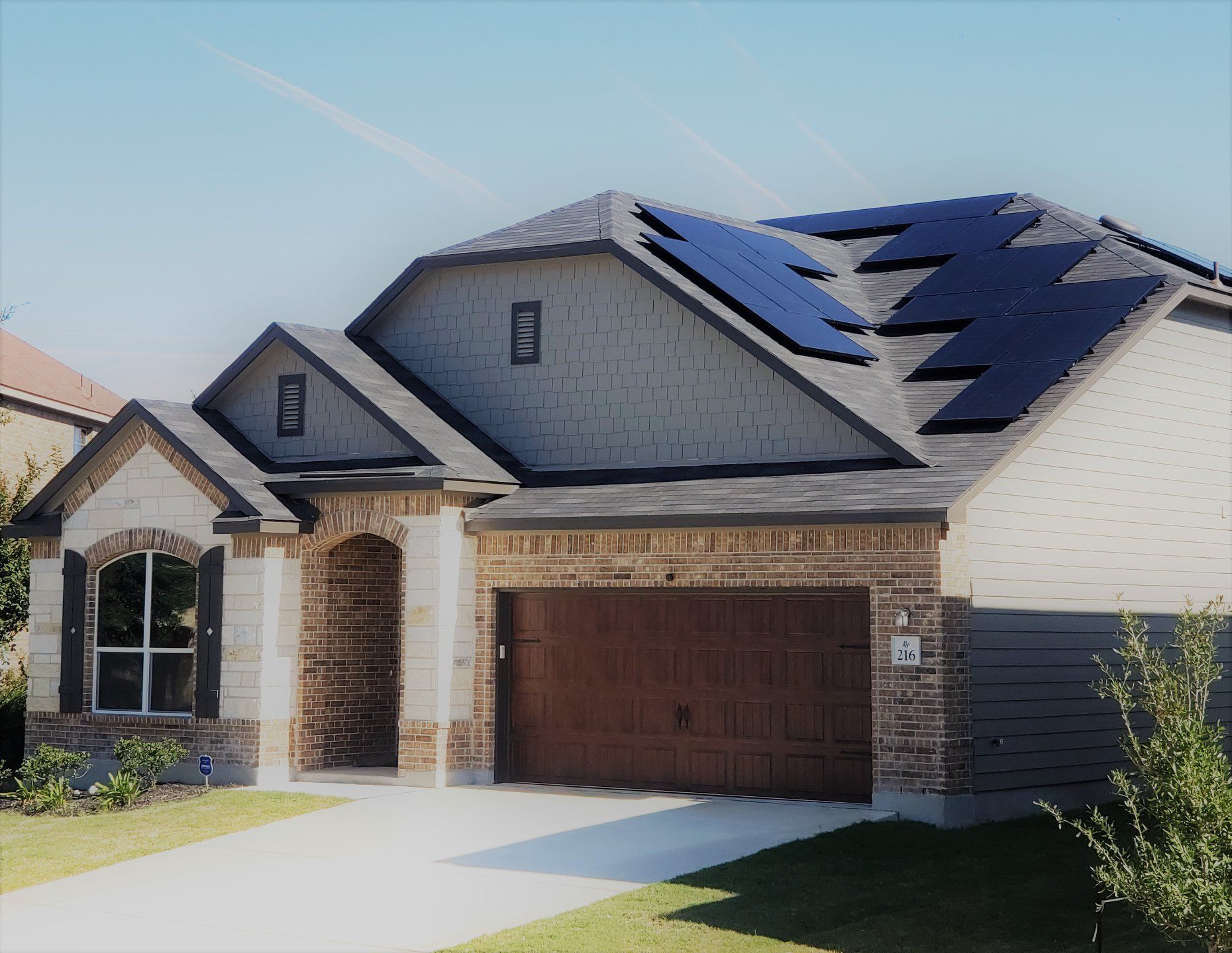 Suburban house with brick garage, stone entry, and dark solar panels on the roof under a clear sky