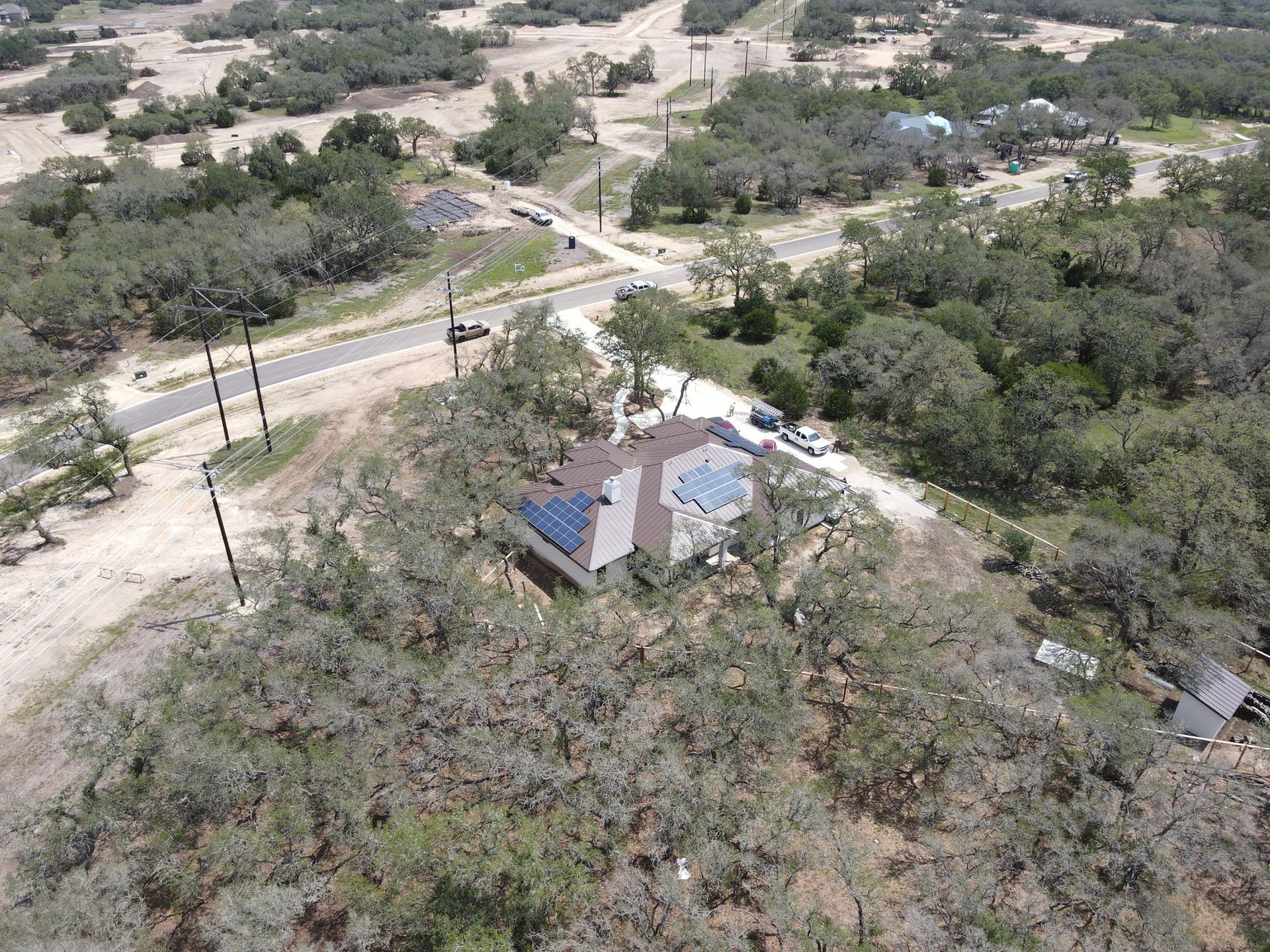 Aerial view of a house with a blue roof beside a road, surrounded by dry trees and scrub.