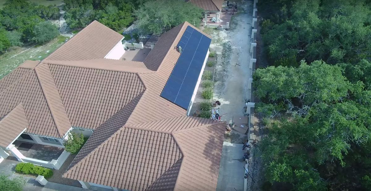 Aerial view of tan-roofed buildings beside a tree-lined road with parked vehicles and solar panels.