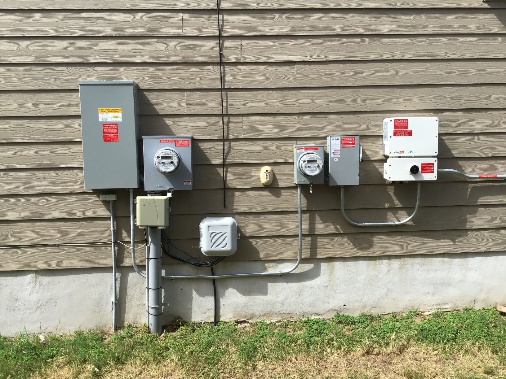 Several outdoor electrical utility boxes and meters mounted on a tan building wall above grass.