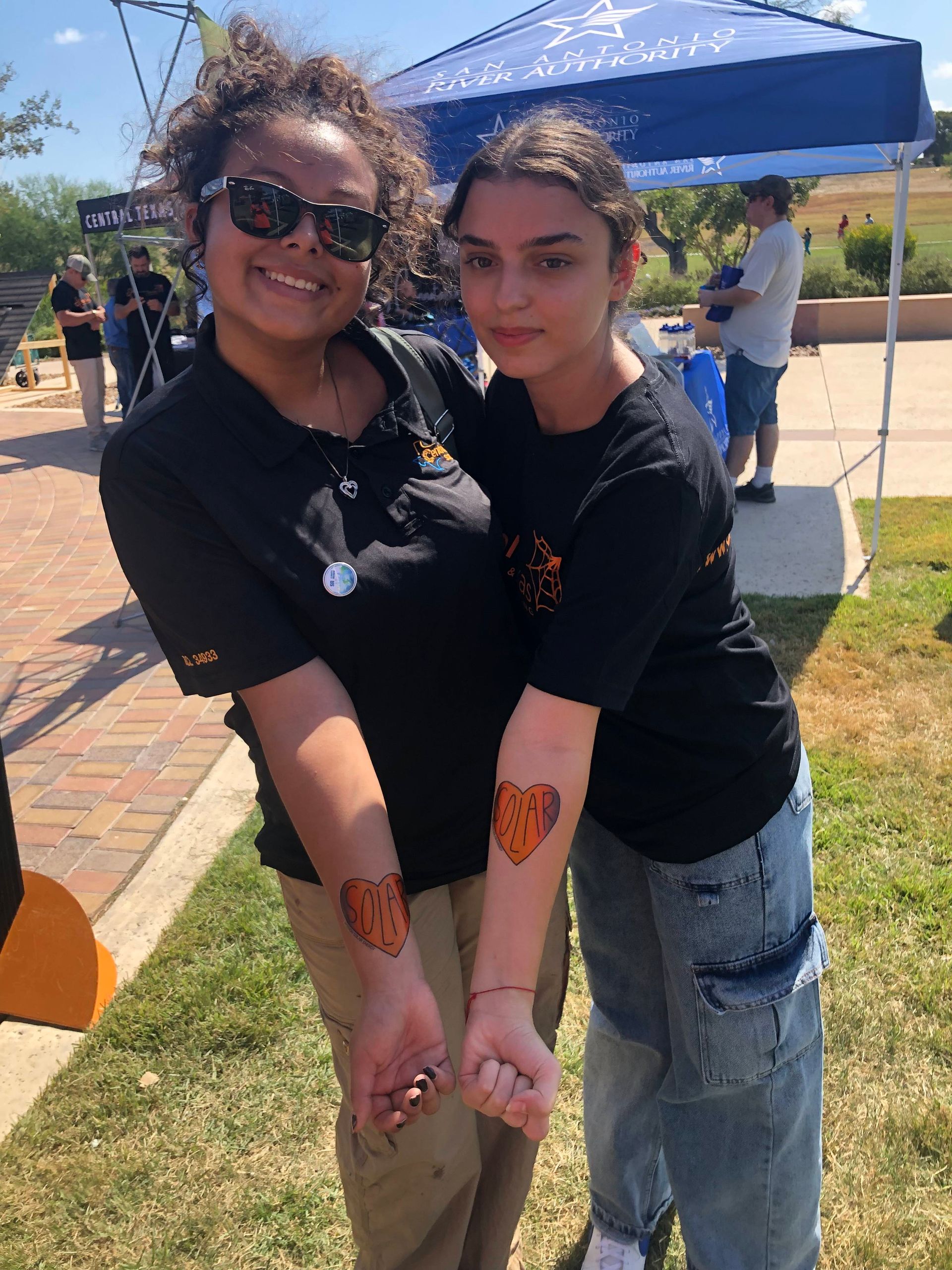 Two people smiling outdoors, wearing black shirts and showing matching orange wrist tattoos.