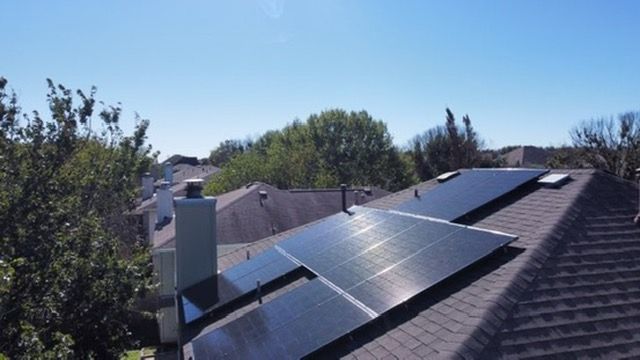 Solar panels on a house roof under a clear blue sky, with trees in the background.