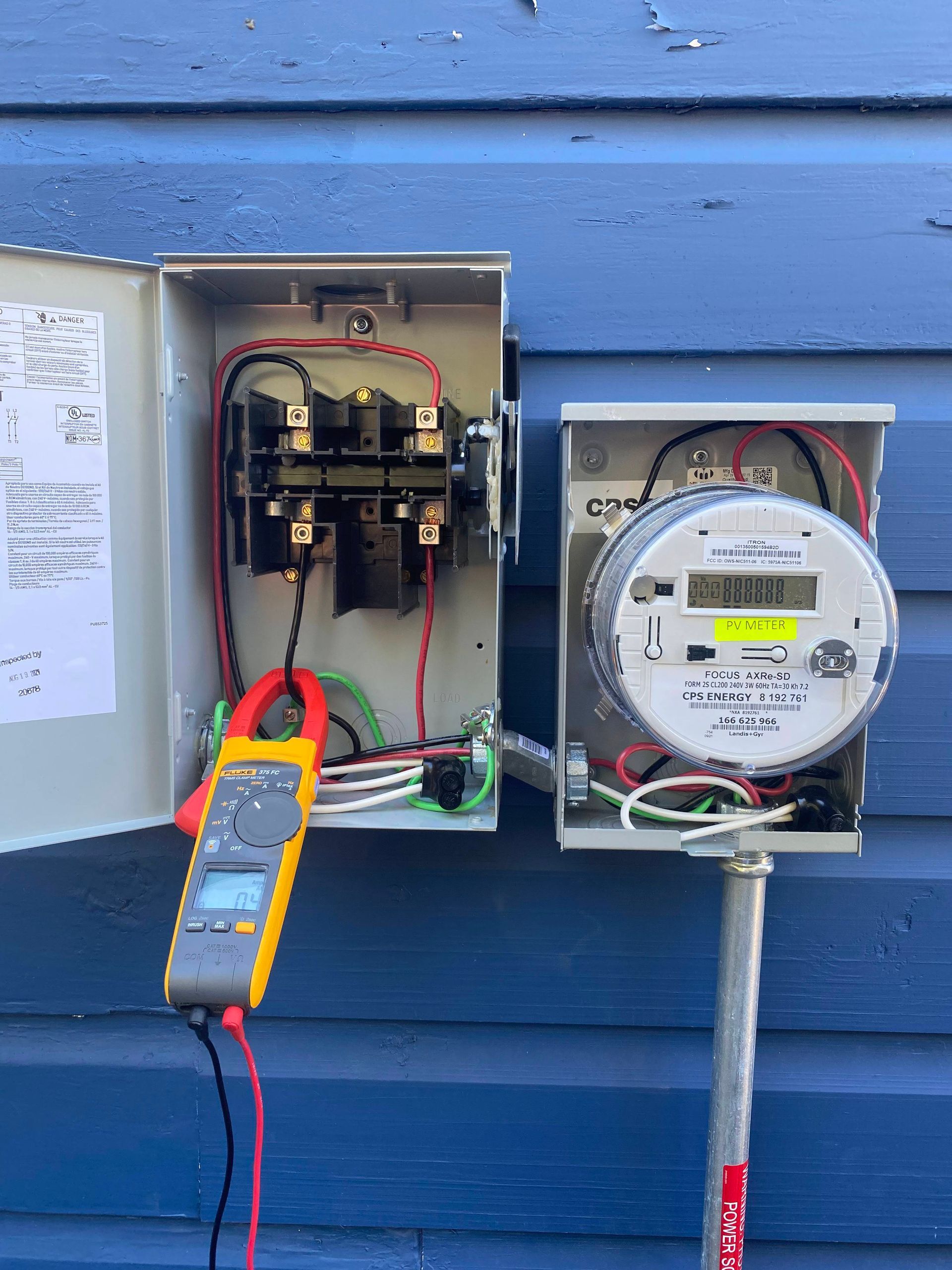 Electric meters and open utility boxes with a multimeter testing wiring on a blue house exterior.
