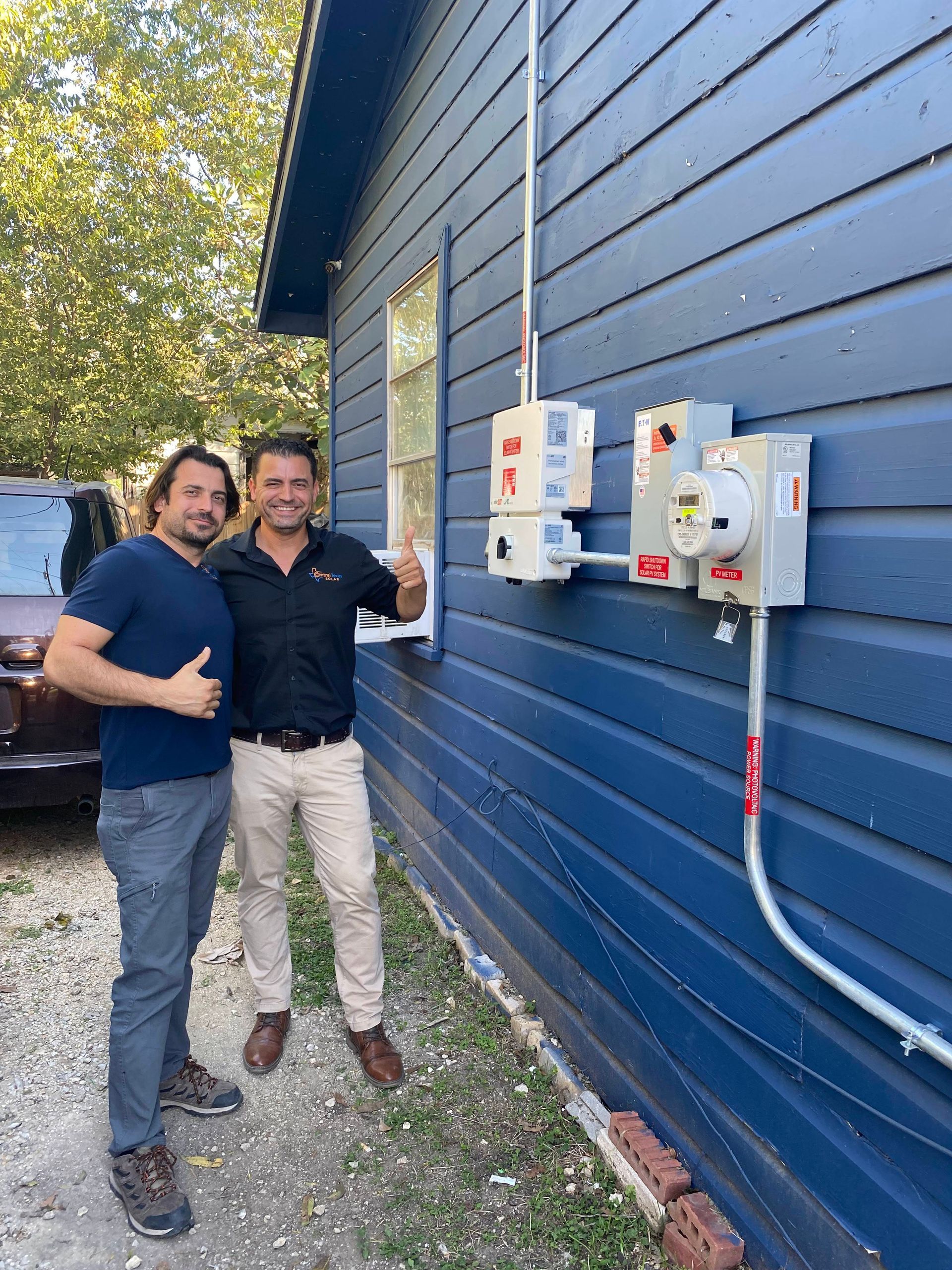 Two people standing beside wall-mounted electrical boxes on a blue building, smiling outdoors.