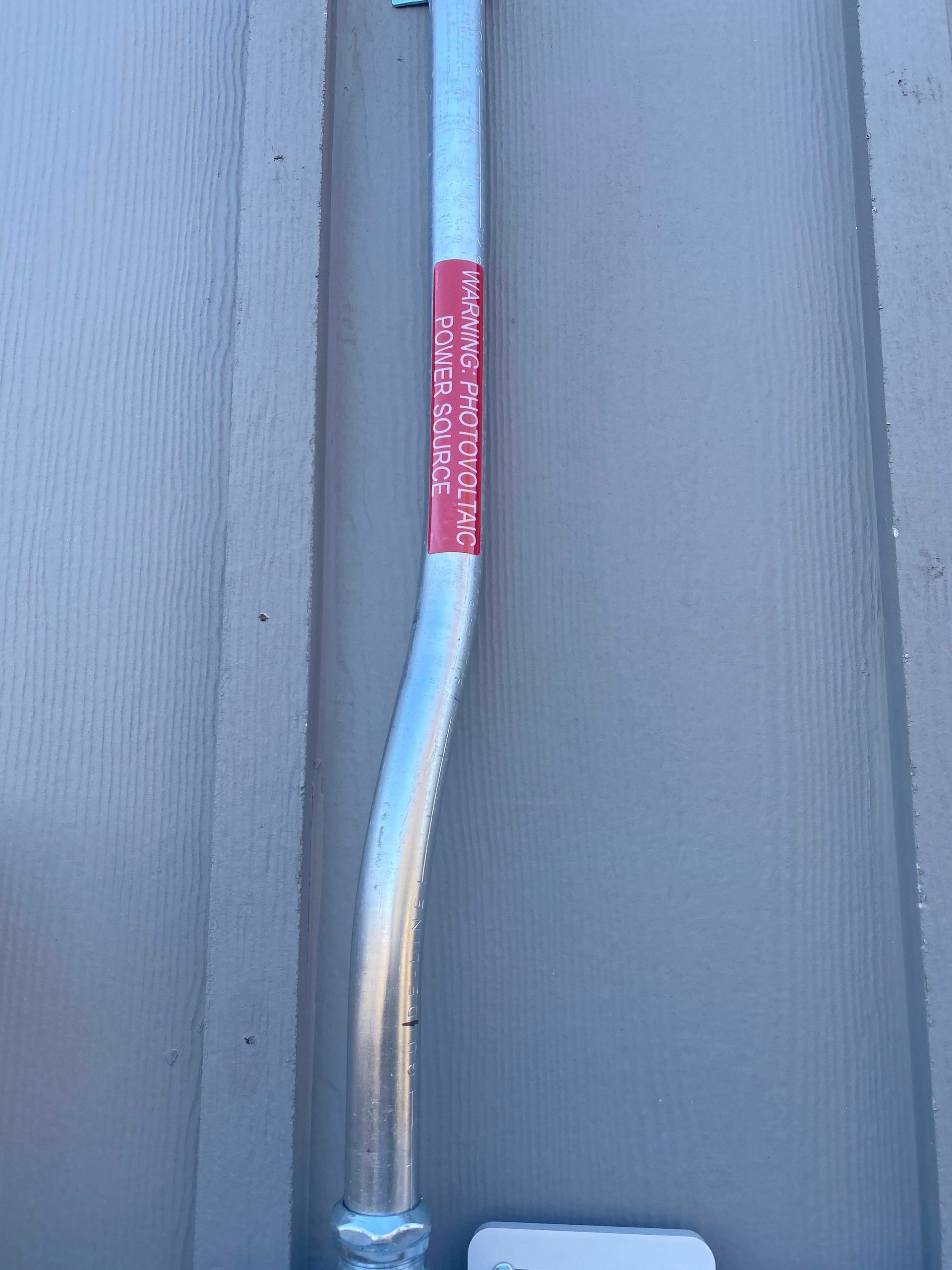 Bent metal pipe with a red label mounted on a gray corrugated wall