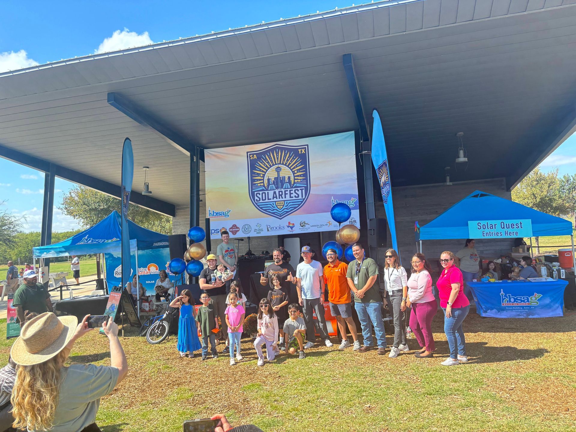 Group posing on a stage under a pavilion with blue balloons and event banner, outdoors on a sunny day