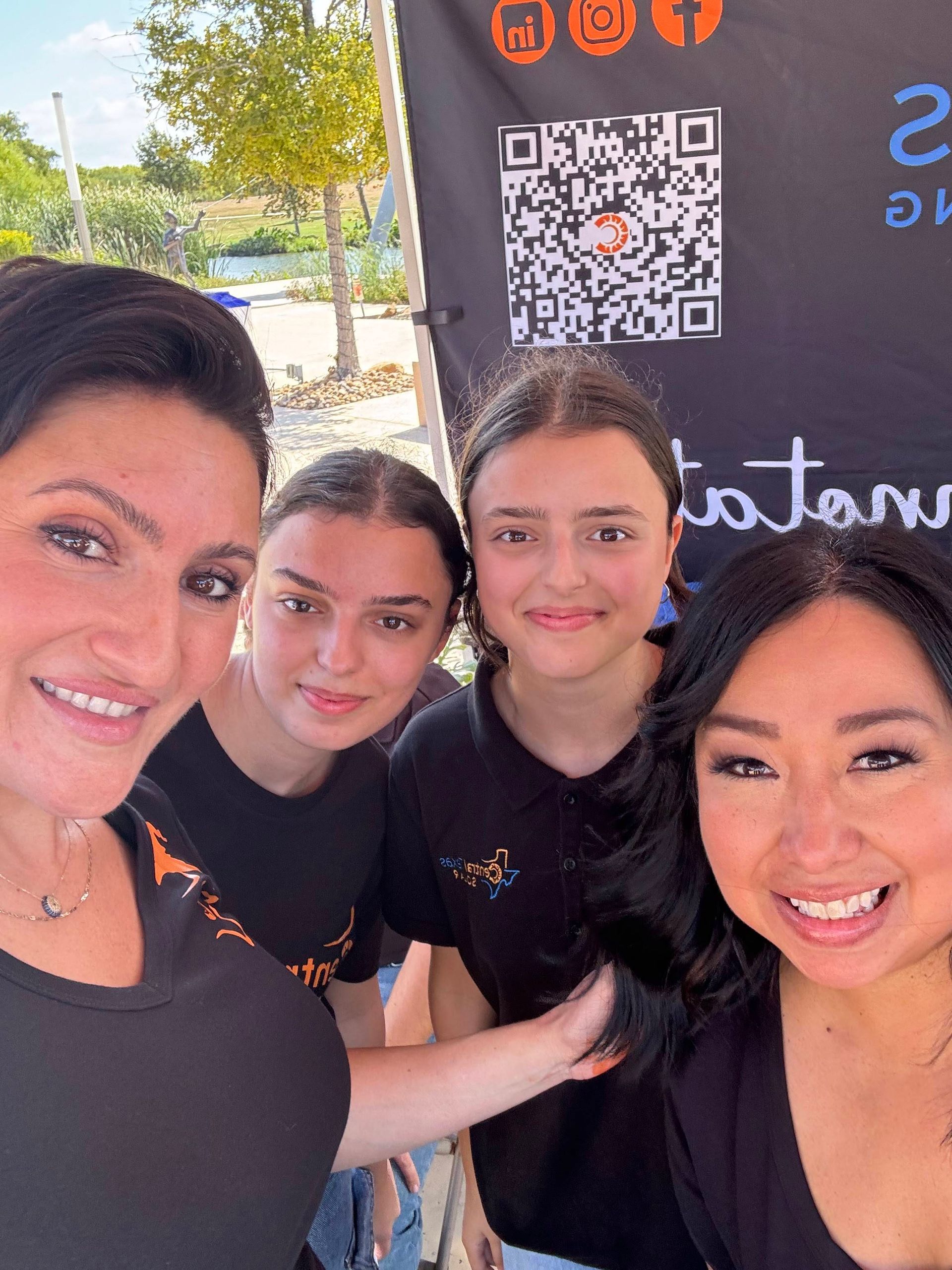 Four smiling people taking a selfie outdoors near a sign with a QR code