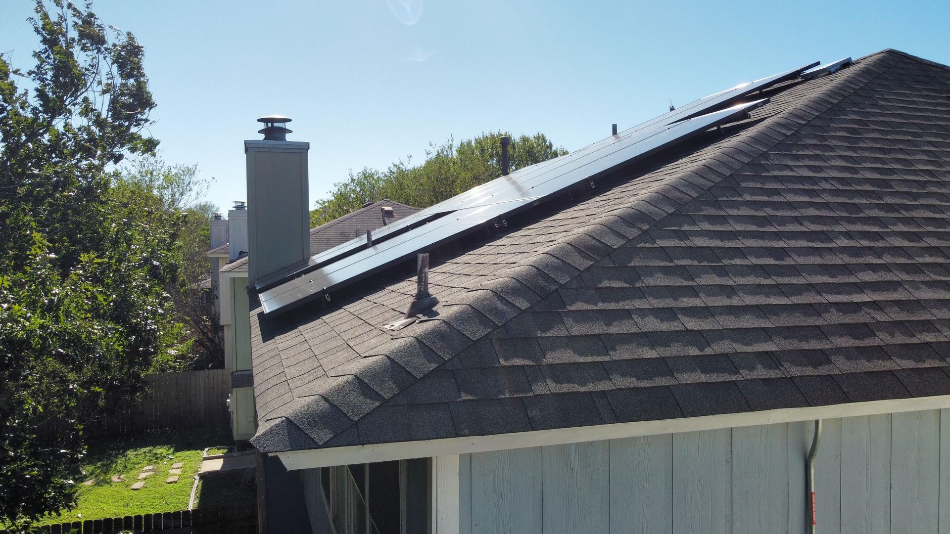 Roof with gray shingles and chimney beside a tree-lined yard on a sunny day