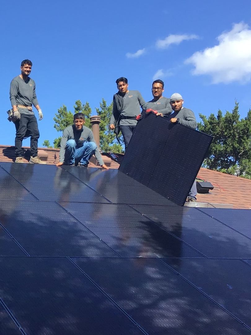 Four workers on a slanted rooftop solar panel installation under a blue sky.