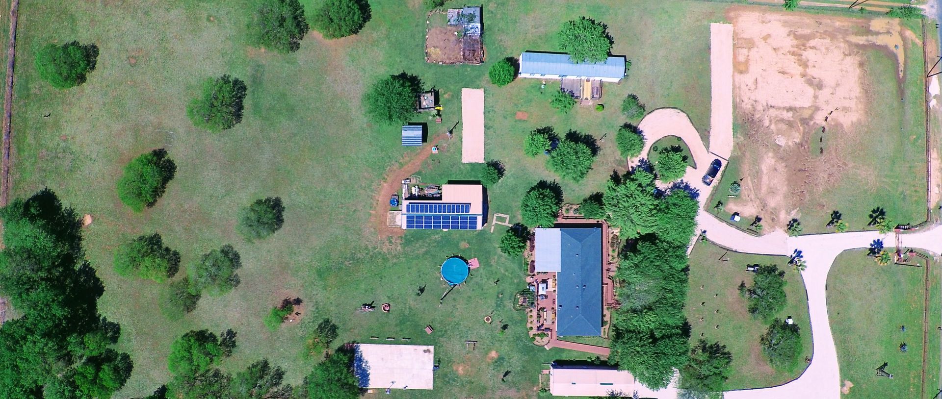 Aerial view of a rural property with several buildings, trees, and a dirt driveway beside open fields
