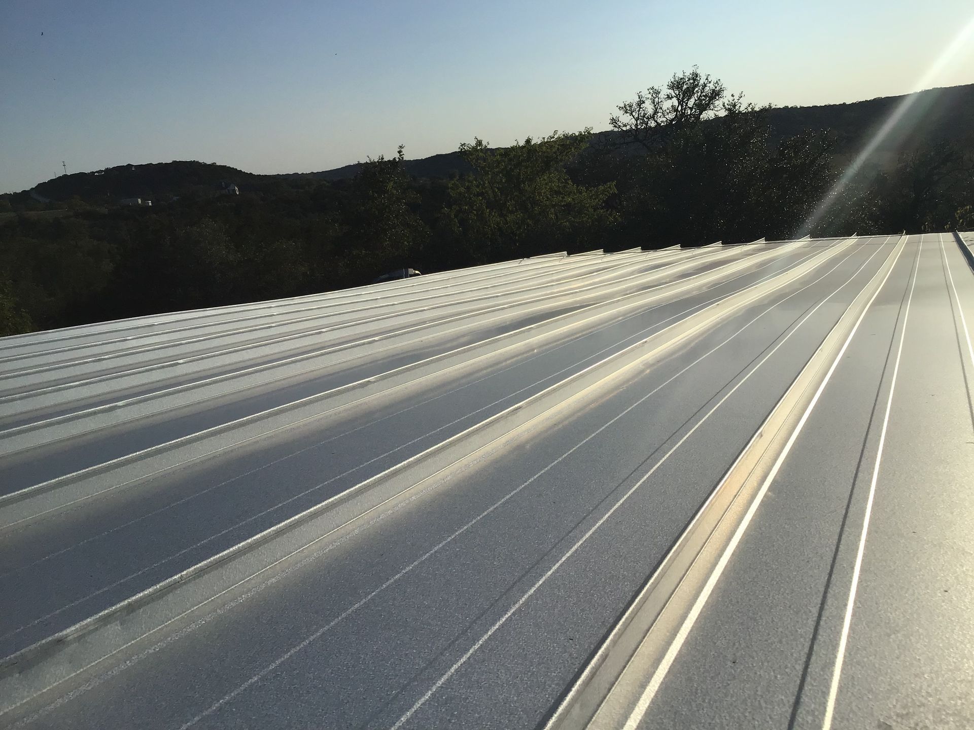 Sunlit white metal roof with long ridges, overlooking a tree-lined landscape at sunset