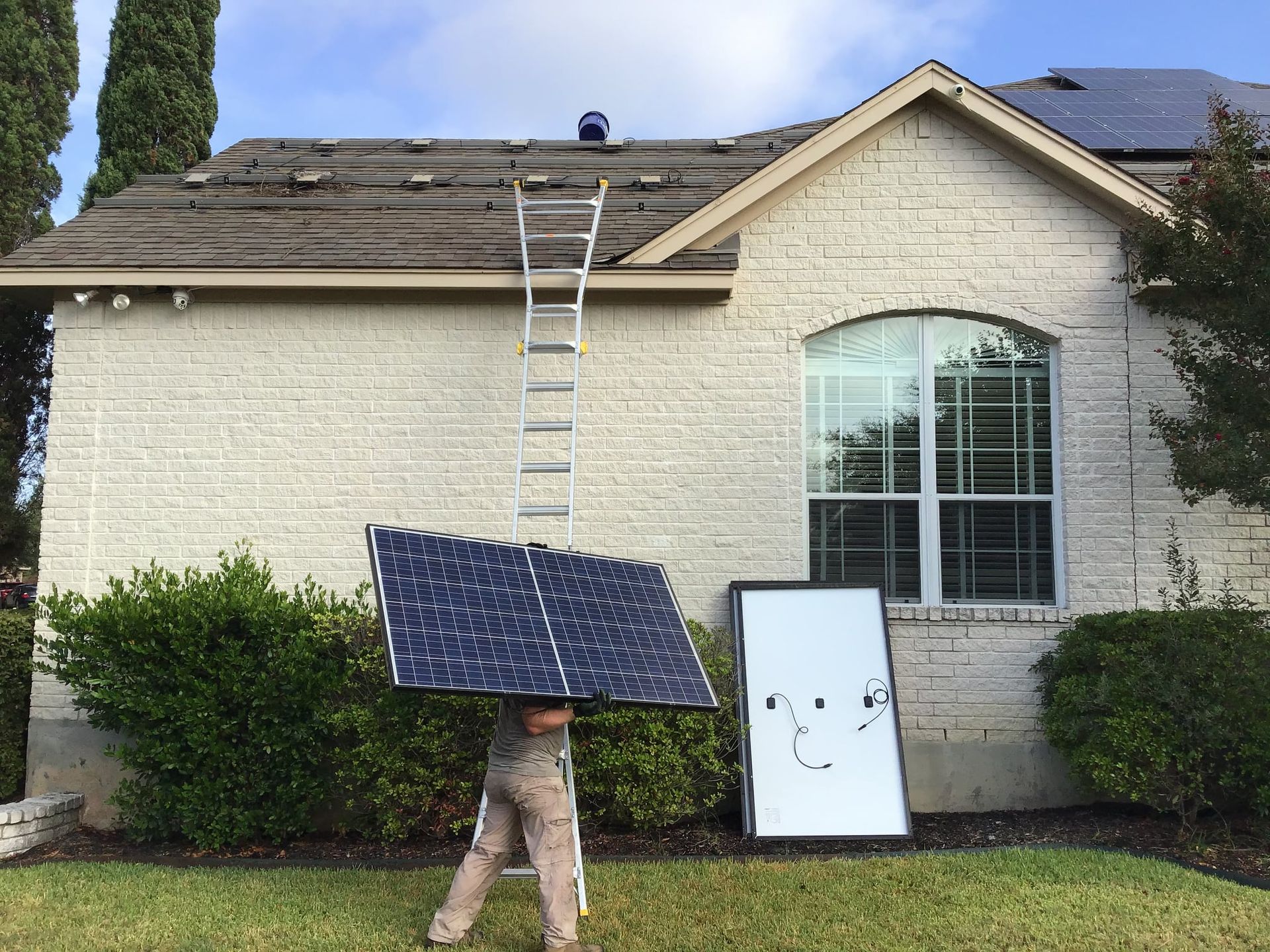 Worker carrying a solar panel beside a ladder and roof-mounted panels on a house