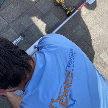 Worker in blue shirt kneeling on a shingled roof, using a tool near a ladder.
