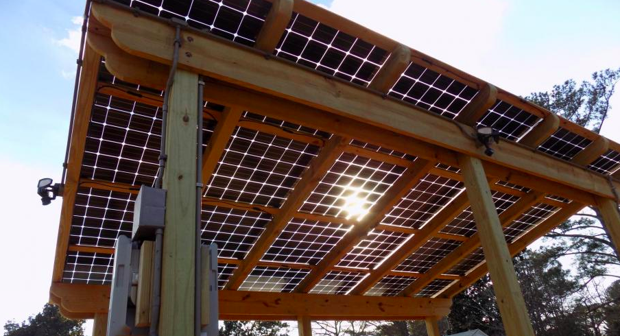 Wooden solar panel canopy with sunlight shining through, viewed from below against a blue sky.