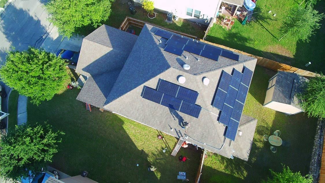 Aerial view of a house with solar panels on the roof, surrounded by trees and a green yard.