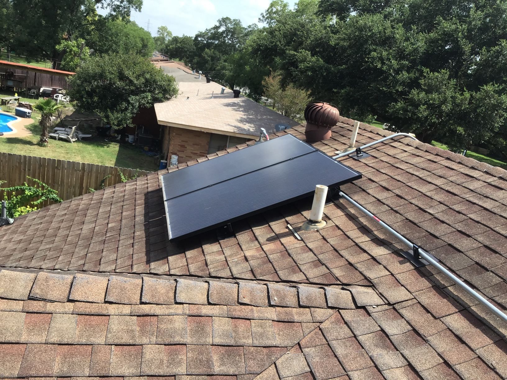 Black solar panel mounted on a brown shingled roof overlooking a wooded neighborhood.
