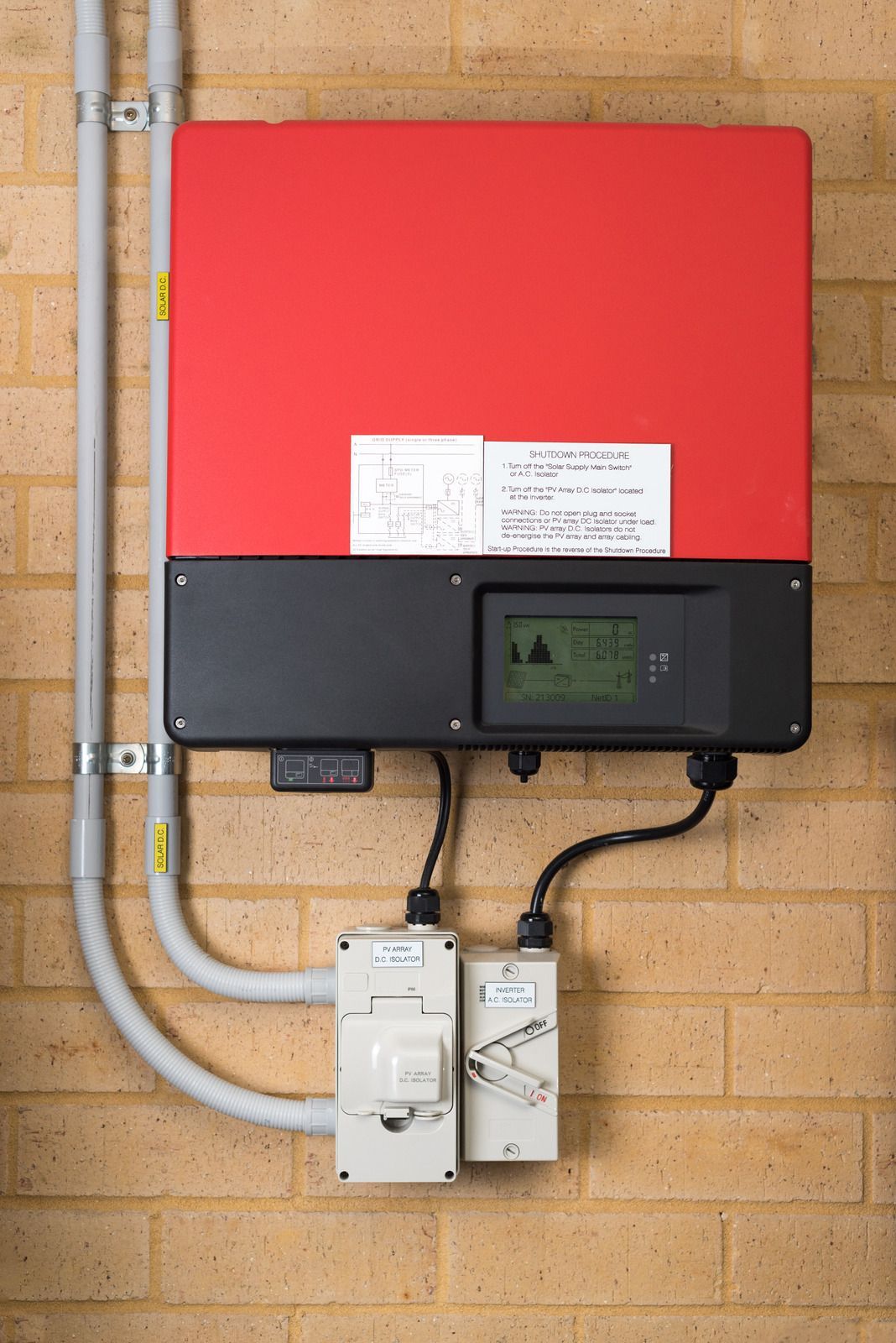 Wall-mounted solar inverter with red and black casing on a brick wall, with connected conduits and control boxes.