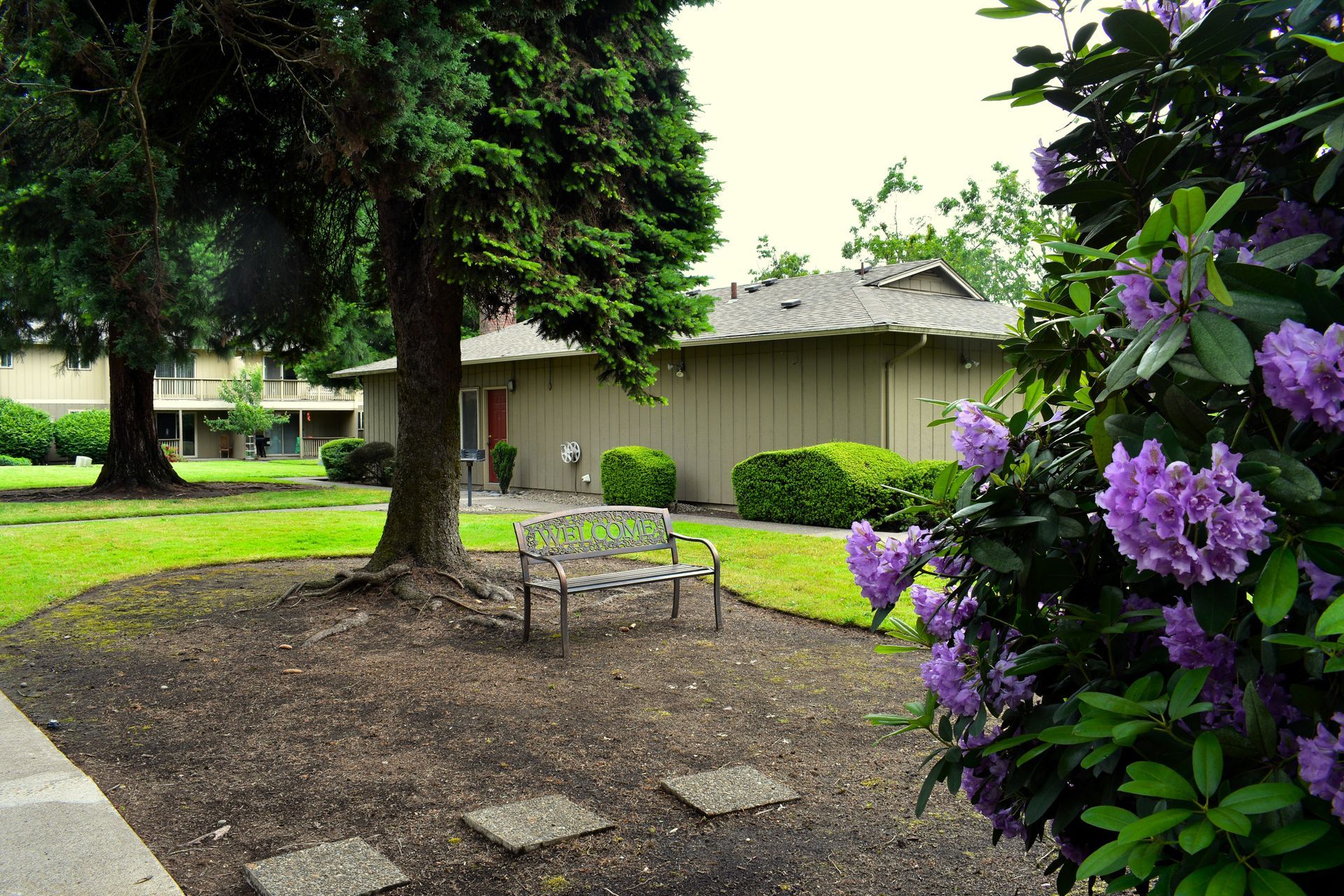 Garden patio with a metal table and chairs, green lawn, house, and purple flowers in the foreground