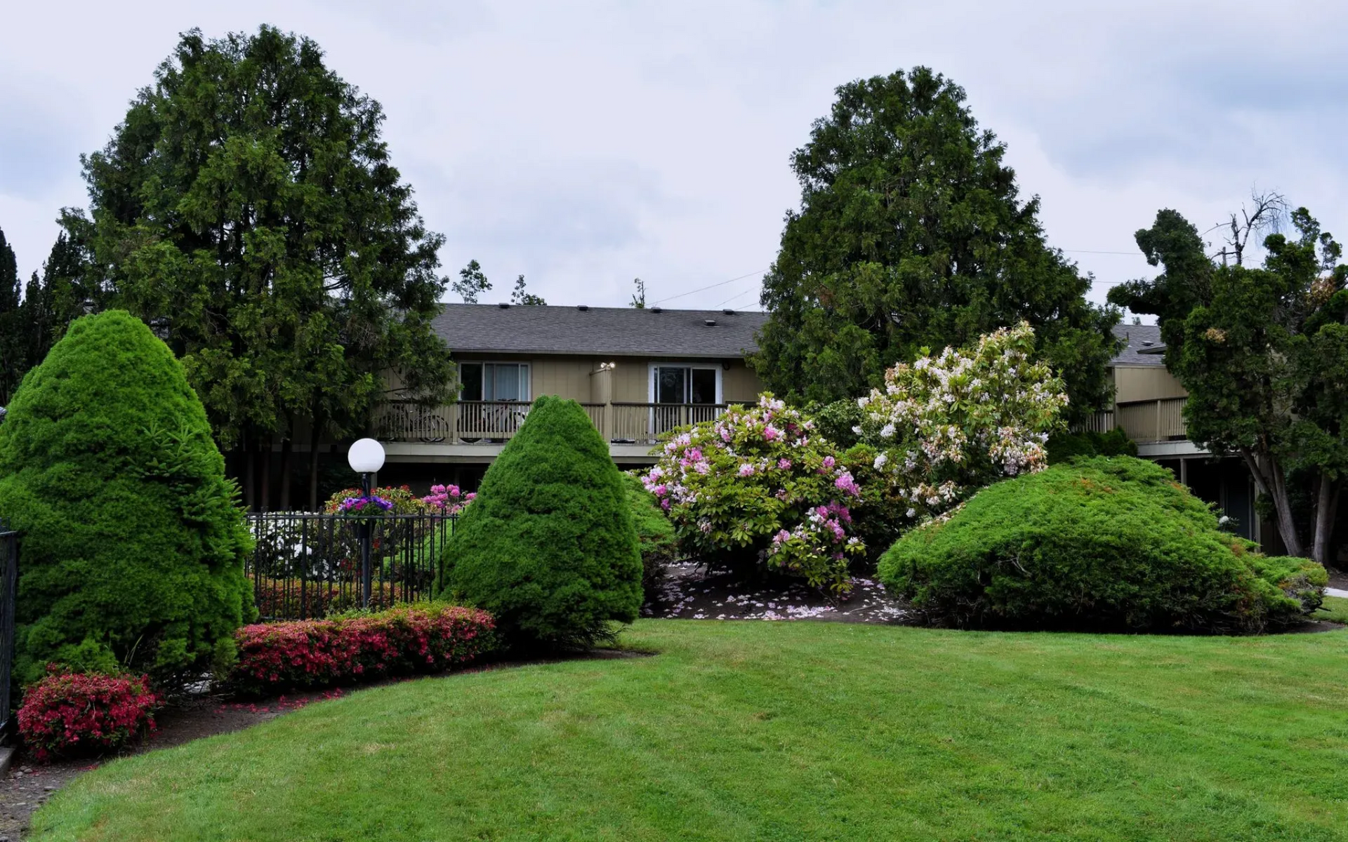 Two-story beige building with balcony, hanging flower baskets, and trees beside a parking lot