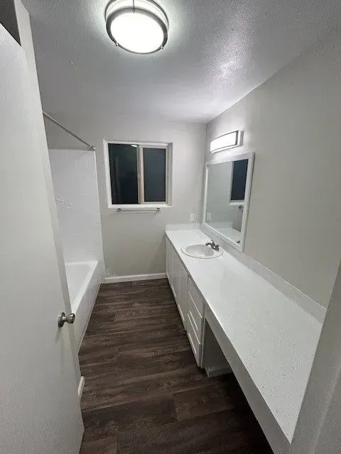 Narrow white bathroom with dark wood floor, wall mirror, sink vanity, window, and ceiling light