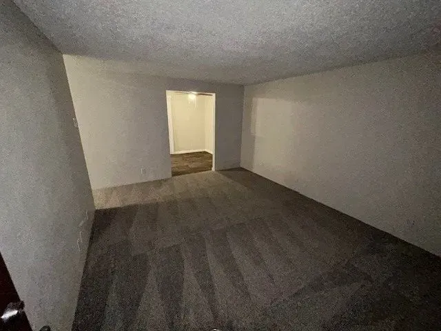 Empty carpeted room with white walls and a doorway opening to a lit hall