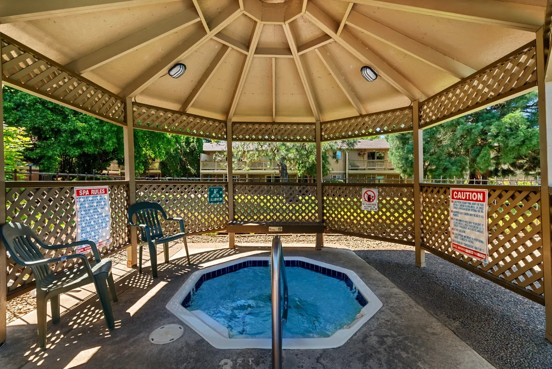 Covered gazebo with a small blue hot tub, benches, and chain-link fencing around a wooded area.