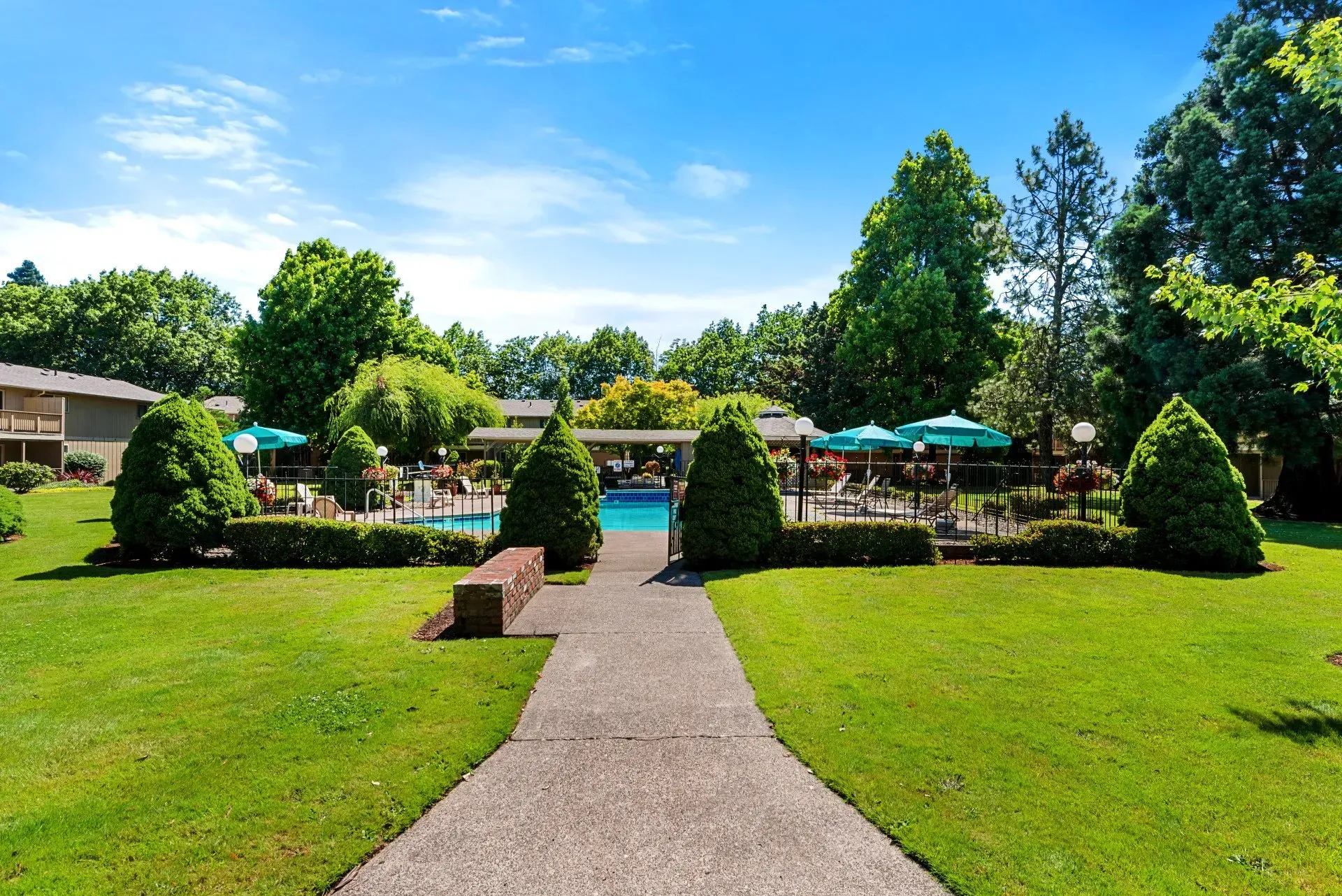 A sunny garden path leading to a patio with umbrellas, trees, and manicured green lawns