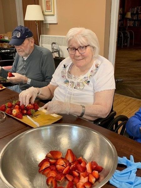 Elderly woman and man slicing strawberries in a well-lit room. Woman in glasses smiles, wearing gloves. Bowl full of cut fruit.