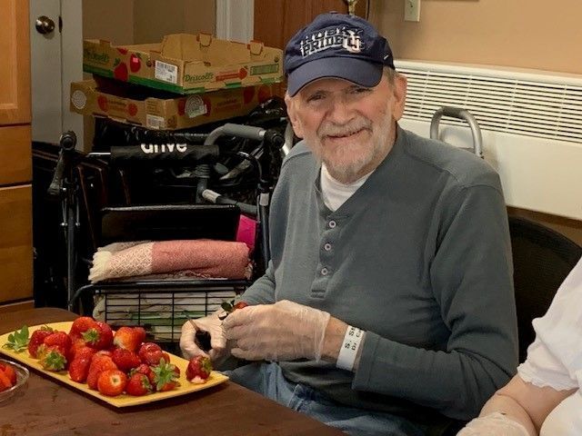 Elderly man in cap and gloves smiles, cutting strawberries at a table.