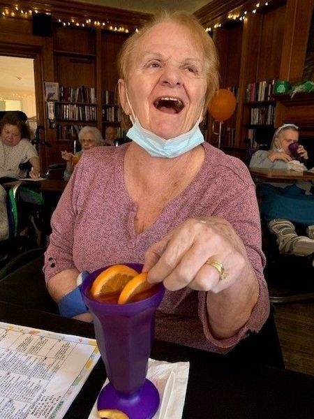 Woman in a senior center, laughing, holding orange slices over a purple drink. Other seniors in background.